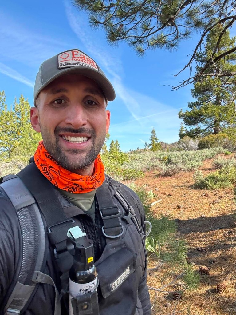 Jason Lombardi smiling in a field cap and orange neck gaiter, wearing a pack and chest harness, with pines and sagebrush behind him